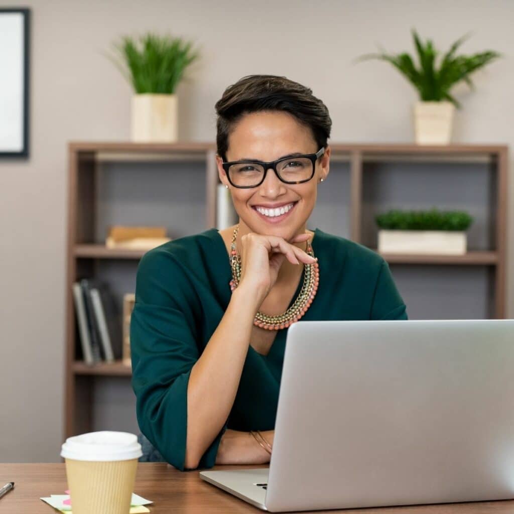 Woman sitting at desk with her hand under her chin smiling, sitting in front of laptop and coffee