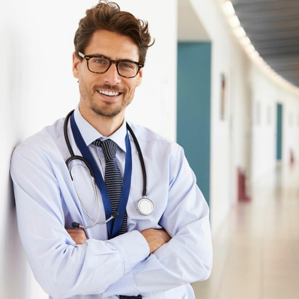 Male doctor in hallway leaning up again wall, arms crossed, smiling
