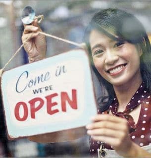 Woman smiling behind a window while hanging a 'Come In We're Open' sign